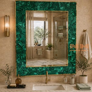 May include: A rectangular wall mirror featuring a wide frame made of green malachite stone with a polished finish and thin gold metal trim. The mirror reflects a bathroom interior with a window, a shower, and a marble countertop vanity.