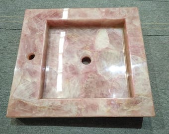 Può includere: Un lavabo quadrato in quarzo rosa con un bacino rettangolare e un foro di scarico. Il lavabo è realizzato in pietra naturale e ha una finitura lucida.