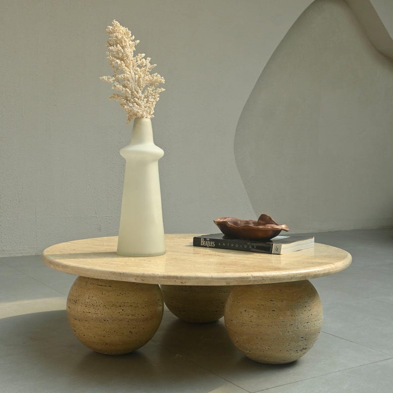 May include: A beige travertine coffee table with three round legs. The table has a white vase with dried flowers and a book on top.