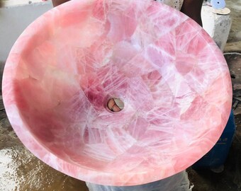 Pode incluir: Uma pia grande, redonda e rosa feita de quartzo rosa com um furo de drenagem no centro. A pia é feita de pedra natural e tem um padrão veteado único.