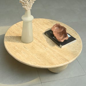 May include: A round travertine coffee table with a unique, rounded base. The table features a light beige color with natural veining and a smooth, polished surface. A white vase with dried branches and a brown ceramic bowl are placed on the table, along with a black book titled "Beatles - The Anthology".