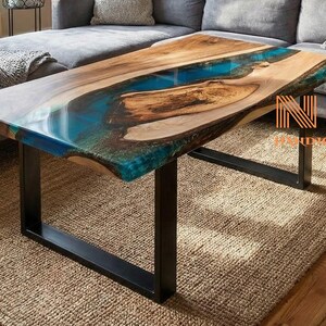 May include: A rectangular coffee table featuring a natural live-edge wood slab with a central translucent blue resin river inlay. The table is supported by a black metal frame and includes the text NM HANDICRAFT in orange.