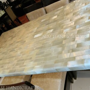 May include: A large rectangular dining table with a white and cream mosaic tile top. The table is made of natural materials and has a rustic, handcrafted look. The text "N.M HANDICRAFTS" is visible on the table.