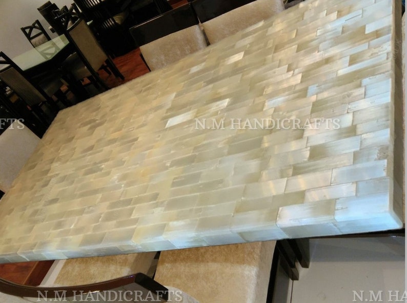 May include: A large rectangular dining table with a white and cream mosaic tile top. The table is made of natural materials and has a rustic, handcrafted look. The text "N.M HANDICRAFTS" is visible on the table.