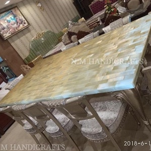 May include: A large dining table with a light blue and beige mosaic top. The table is surrounded by chairs with silver frames and gray velvet seats. The table is made of natural stone and has a unique, handcrafted look. The text "N.M HANDICRAFTS" is visible on the table.