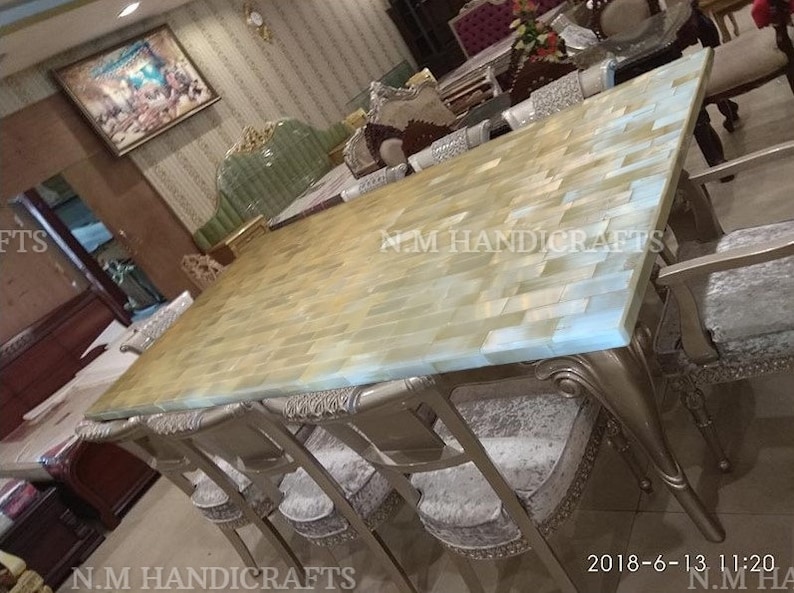 May include: A large dining table with a light blue and beige mosaic top. The table is surrounded by chairs with silver frames and gray velvet seats. The table is made of natural stone and has a unique, handcrafted look. The text "N.M HANDICRAFTS" is visible on the table.