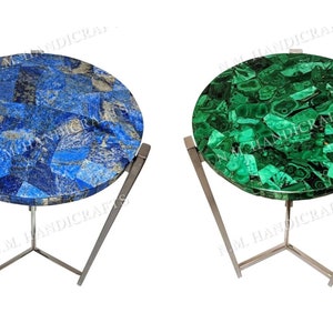 May include: Two round side tables with a unique design. The tabletops are made of a mosaic of blue and green gemstone pieces. The tables have a silver metal base with a tripod design.