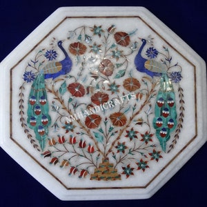 May include: Octagonal white marble table top with intricate inlaid peacock and floral design. The design features peacocks with blue and green feathers, red and orange flowers, and green leaves. The table top is inlaid with various colored stones, including turquoise, lapis lazuli, and mother of pearl. The table top is signed "NMHANDICRAFTS".