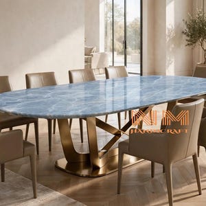 Puede incluir: Una mesa de comedor ovalada con una superficie de mármol azul pulido con vetas blancas y una base de metal dorado con patas entrecruzadas, rodeada de ocho sillas tapizadas en color beige con reposabrazos. La mesa incluye el texto NM HANDCRAFT.