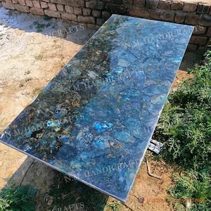 Op de afbeelding: Een groot, rechthoekig tafelblad gemaakt van blauw en groen labradoritsteen. De steen is gepolijst en heeft een reflecterend oppervlak. Het tafelblad is op een metalen frame geplaatst.