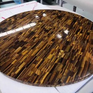 May include: Round table top made of tiger's eye stone. The surface is a mosaic of small, rectangular pieces of polished tiger's eye, creating a shimmering, golden brown effect.