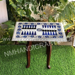 May include: A white marble backgammon game board with blue and gold inlay designs. The board is set up for play with black and white playing pieces. The board is on a wooden table with turned legs.