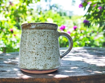 May include: A speckled light gray ceramic mug with a brown rim and handle. The mug is sitting on a weathered wooden surface.