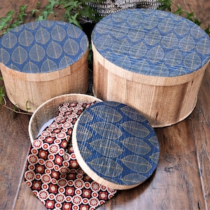 Peut inclure: Trois boîtes rondes en bois avec des couvercles à motifs de feuilles bleues et dorées. Les boîtes sont empilées les unes sur les autres. La boîte du haut est ouverte et un tissu à motifs rouge et blanc est à l'intérieur.