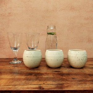 May include: Three white ceramic cups with a speckled texture and two clear glass wine glasses on a wooden table. A clear glass bottle with a green leaf inside is behind the cups.