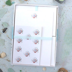 May include: A white box with a blue ribbon contains a set of stationery with a seashell design. The stationery features a seashell with the letter "A" in blue on a pink background. There are 10 seashells with the letter "A" on the stationery.