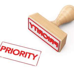 May include: A wooden rubber stamp with a red "PRIORITY" imprint. The stamp is on a white surface and has been used to create a red "PRIORITY" imprint on the surface.