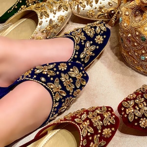 May include: A close-up of a pair of red, blue, and black velvet shoes with gold embellishments. The shoes are decorated with intricate floral patterns and sparkling rhinestones. The shoes are worn by a person's feet, and the brand name "Sandals House" is visible on the sole of one of the shoes.