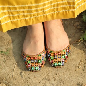 May include: A pair of black flat shoes with colorful embroidered embellishments. The shoes have a pointed toe and are decorated with small, round mirrors and sequins.