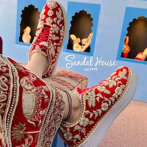 May include: Red sneakers with detailed gold and silver floral embroidery. The shoes have white soles and red laces. The text "Sandal House est 1979" is visible. The shoes are part of a traditional or cultural outfit.