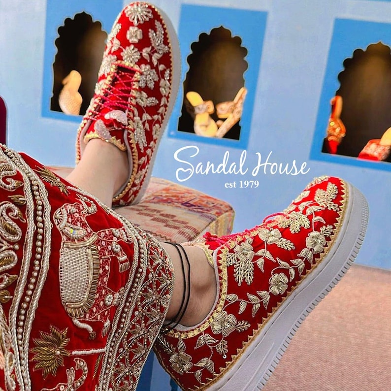 May include: Red sneakers with detailed gold and silver floral embroidery. The shoes have white soles and red laces. The text "Sandal House est 1979" is visible. The shoes are part of a traditional or cultural outfit.