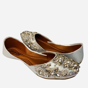 May include: A pair of silver embellished flat shoes with a pointed toe. The shoes are decorated with silver and clear rhinestones. The shoes have a brown leather insole and a silver fabric upper. The brand name "Sandal House" is printed on the insole.
