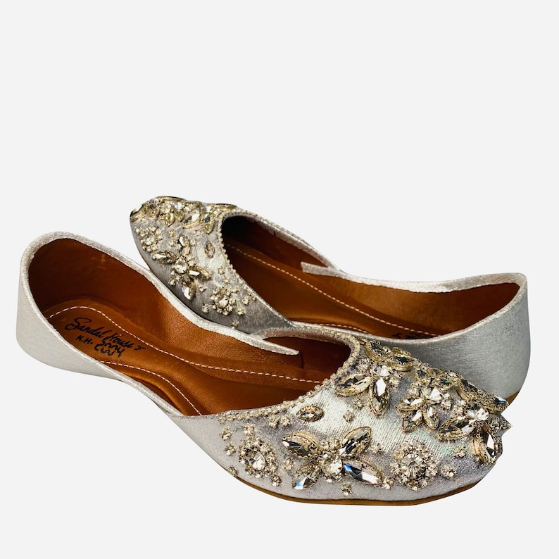 May include: A pair of silver embellished flat shoes with a pointed toe. The shoes are decorated with silver and clear rhinestones. The shoes have a brown leather insole and a silver fabric upper. The brand name "Sandal House" is printed on the insole.