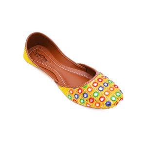 May include: Yellow embroidered flat shoes with colorful round mirror embellishments. The shoes have a brown leather insole and a pointed toe.