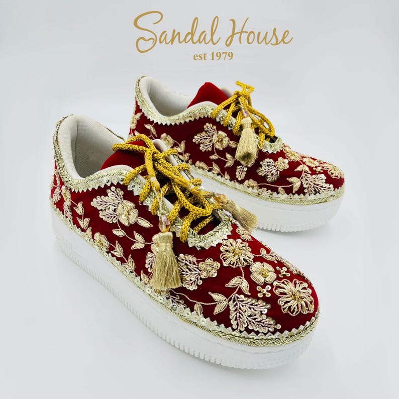 May include: Red sneakers with gold floral embroidery, white soles, and gold tassel laces. The shoes feature the text "Sandal House est 1979". The design is intricate and the colour scheme is striking. The shoes are a unique fashion item.