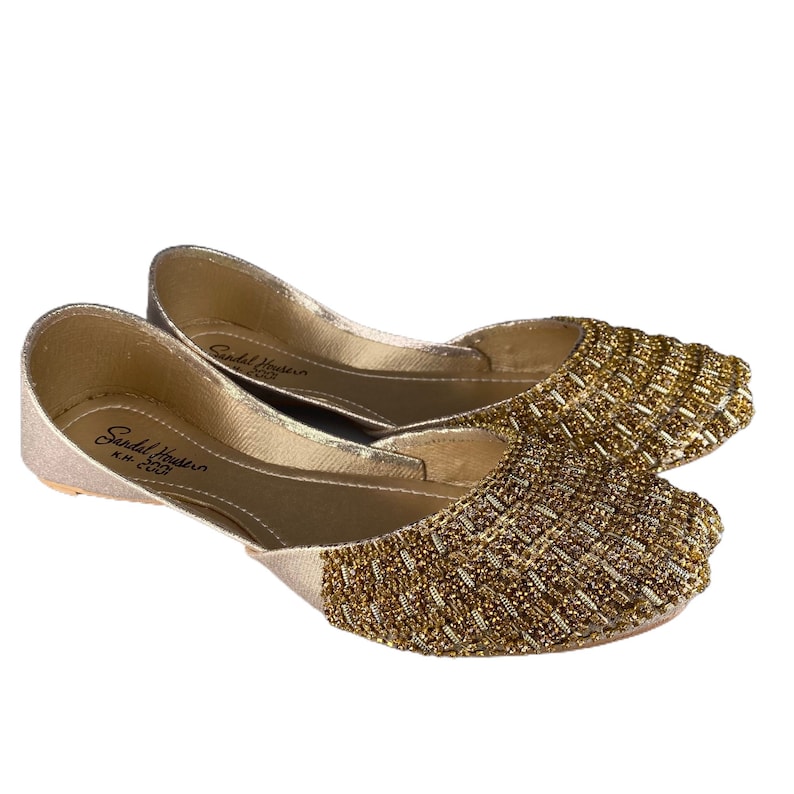 Puede incluir: Un par de zapatos planos dorados con un dise&ntilde;o de cuentas. Los zapatos tienen una punta puntiaguda y un tac&oacute;n bajo. El nombre de la marca "Sandal House" es visible en la plantilla.