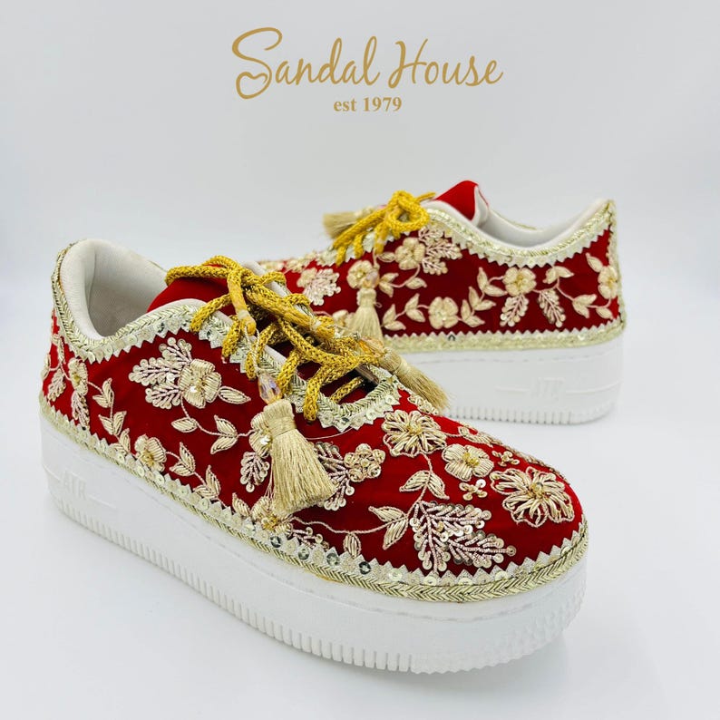 May include: Red sneakers with gold floral embroidery and white soles. The shoes feature gold laces with tassels and are trimmed with gold sequins. The brand name "Sandal House" is visible in gold script.