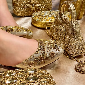 May include: A pair of gold sparkly flats with a matching gold clutch purse. The purse has a chain strap and is decorated with rhinestones. The flats are also decorated with rhinestones and have a pointed toe.