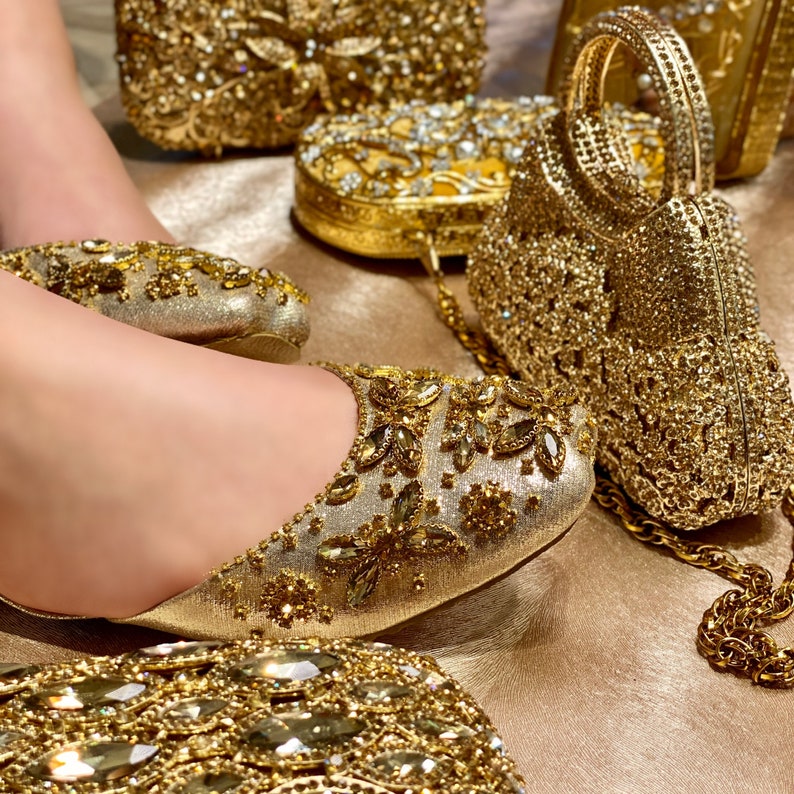 May include: A pair of gold sparkly flats with a matching gold clutch purse. The purse has a chain strap and is decorated with rhinestones. The flats are also decorated with rhinestones and have a pointed toe.