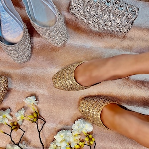 Puede incluir: Un par de zapatos planos de punta con adornos dorados. Los zapatos est&aacute;n sobre una superficie marr&oacute;n claro con un bolso de mano decorativo de cuentas plateadas.