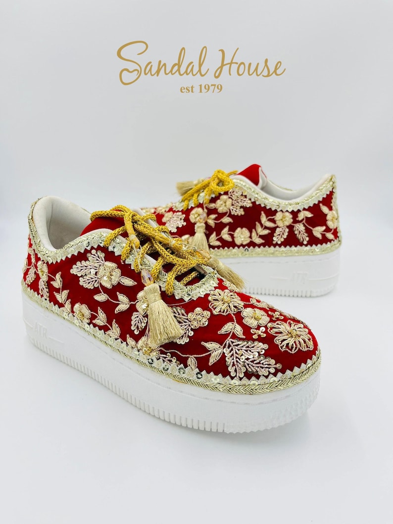 May include: Red sneakers with gold floral embroidery and tassels. The shoes have white soles and gold trim. The text "Sandal House est 1979" is in gold script above the shoes.