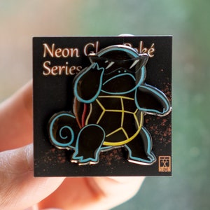 May include: A black and yellow enamel pin featuring a cartoon turtle wearing sunglasses. The pin is on a black card with the text "Neon Classic Series" and a logo.