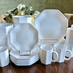 May include: A set of white octagonal dinnerware, including plates, bowls, and mugs. The set is arranged on a wooden table with a floral centerpiece in the background.