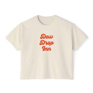 以下が含まれることがあります： レトロなオレンジ色のフォントで「Dew Drop Inn」と書かれたクリーム色のTシャツ。 シャツはクルーネックと半袖です。 カジュアルなTシャツは、綿または同様の生地で作られている可能性があります。