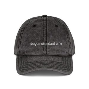 May include: A distressed black baseball cap with a curved brim. The cap features the phrase "dragon standard time" embroidered in white across the front. The hat has a classic six-panel design and an adjustable strap.
