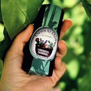 Può includere: Un deodorante in stick cilindrico nero con un'etichetta verde che riporta il testo Dawn to Dusk Alchemy e Lemongrass Deodorant. L'etichetta mostra l'illustrazione di un mortaio scuro con pestello riempito di lavanda, erbe e fiori bianchi.
