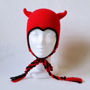 Puede incluir: Un gorro de ganchillo rojo brillante con dos cuernos puntiagudos en la parte superior. El gorro tiene un borde negro a lo largo de la abertura facial y dos largas tiras trenzadas de hilo rojo y negro que cuelgan a los lados, terminadas en borlas.