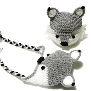 May include: A gray and white crocheted wolf hat with black eyes and nose. The hat has two long braided tails.