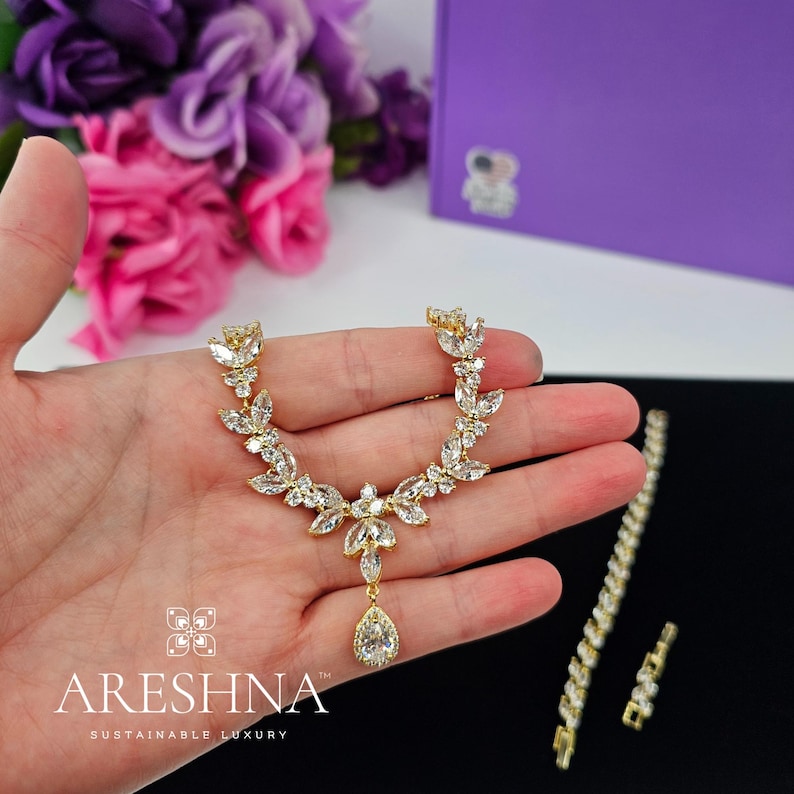 Hand holding The Aurora bridal necklace and earrings set by Areshna™, 18K gold floral jewelry with teardrop crystals, captured in natural light.