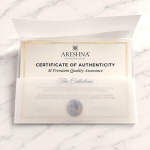 Macro shot of the official Areshna™ Certificate of Authenticity featuring elegant gold foil borders and embossed branding on premium cream textured paper. The document is layered behind a folded translucent vellum sleeve on a white marble background, featuring a handcrafted wax seal, evoking a high-end sustainable luxury unboxing experience included with every order.