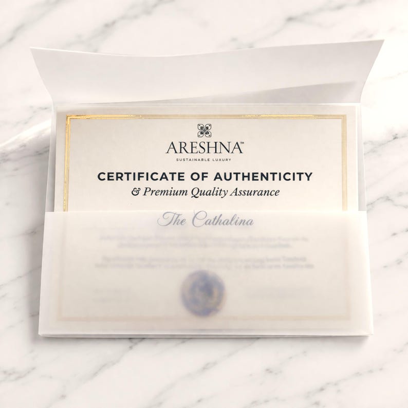 Macro shot of the official Areshna™ Certificate of Authenticity featuring elegant gold foil borders and embossed branding on premium cream textured paper. The document is layered behind a folded translucent vellum sleeve on a white marble background, featuring a handcrafted wax seal, evoking a high-end sustainable luxury unboxing experience included with every order.