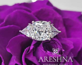 May include: A silver ring featuring a large, cushion-cut diamond in the centre and two smaller, triangular diamonds on either side. The ring is displayed on a bed of purple rose petals.
