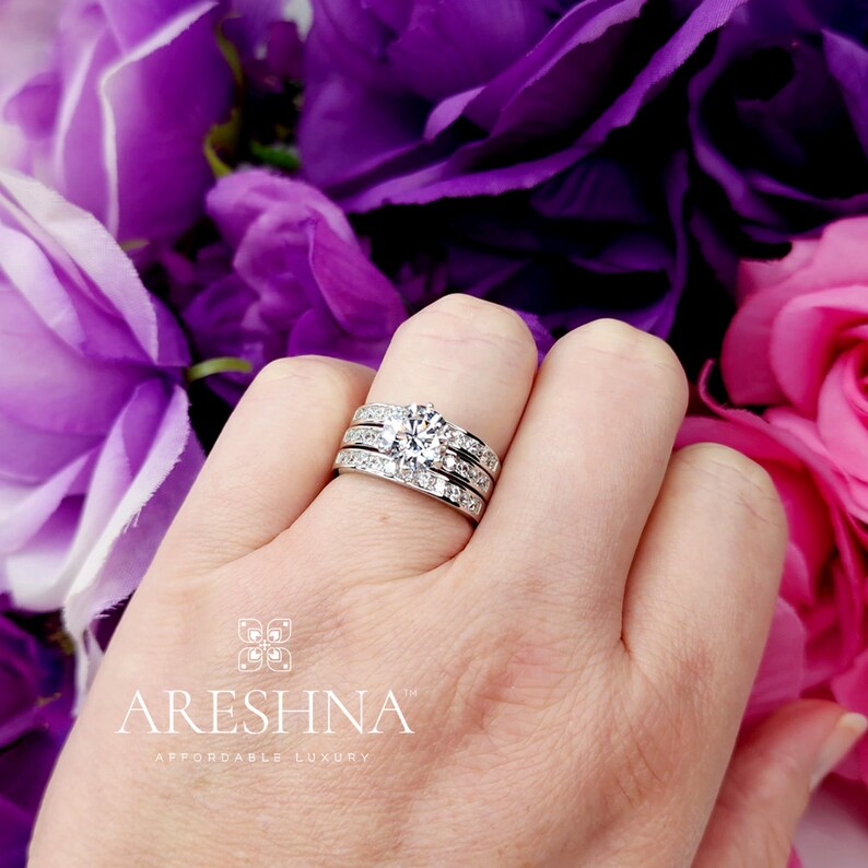 Puede incluir: Un juego de anillos de boda de tres piezas con una piedra central grande, redonda y clara y piedras m&aacute;s peque&ntilde;as en la banda. El anillo est&aacute; en una mano con flores moradas y rosas en el fondo.