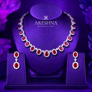 May include: A necklace and earring set featuring oval-cut red stones surrounded by small white stones in a silver-toned metal setting. The necklace is displayed on a stand with the text ARESHNA SUSTAINABLE LUXURY printed in white above the jewelry.