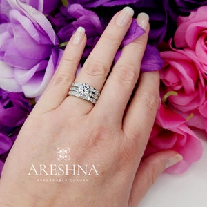 Puede incluir: Un juego de anillos de boda de tres piezas con una piedra central grande y redonda y dos bandas m&aacute;s peque&ntilde;as con diamantes engastados en pav&eacute;. Los anillos est&aacute;n en una mano con flores rosas y moradas en el fondo.
