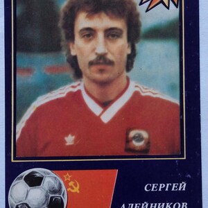 May include: A rectangular sports trading card featuring a portrait of a man with brown hair and a mustache wearing a red jersey. The card includes the text ZVEZDY MIROVOGO FUTBOLA, a star graphic, a soccer ball, the USSR flag, and the name SERGEI ALEINIKOV.